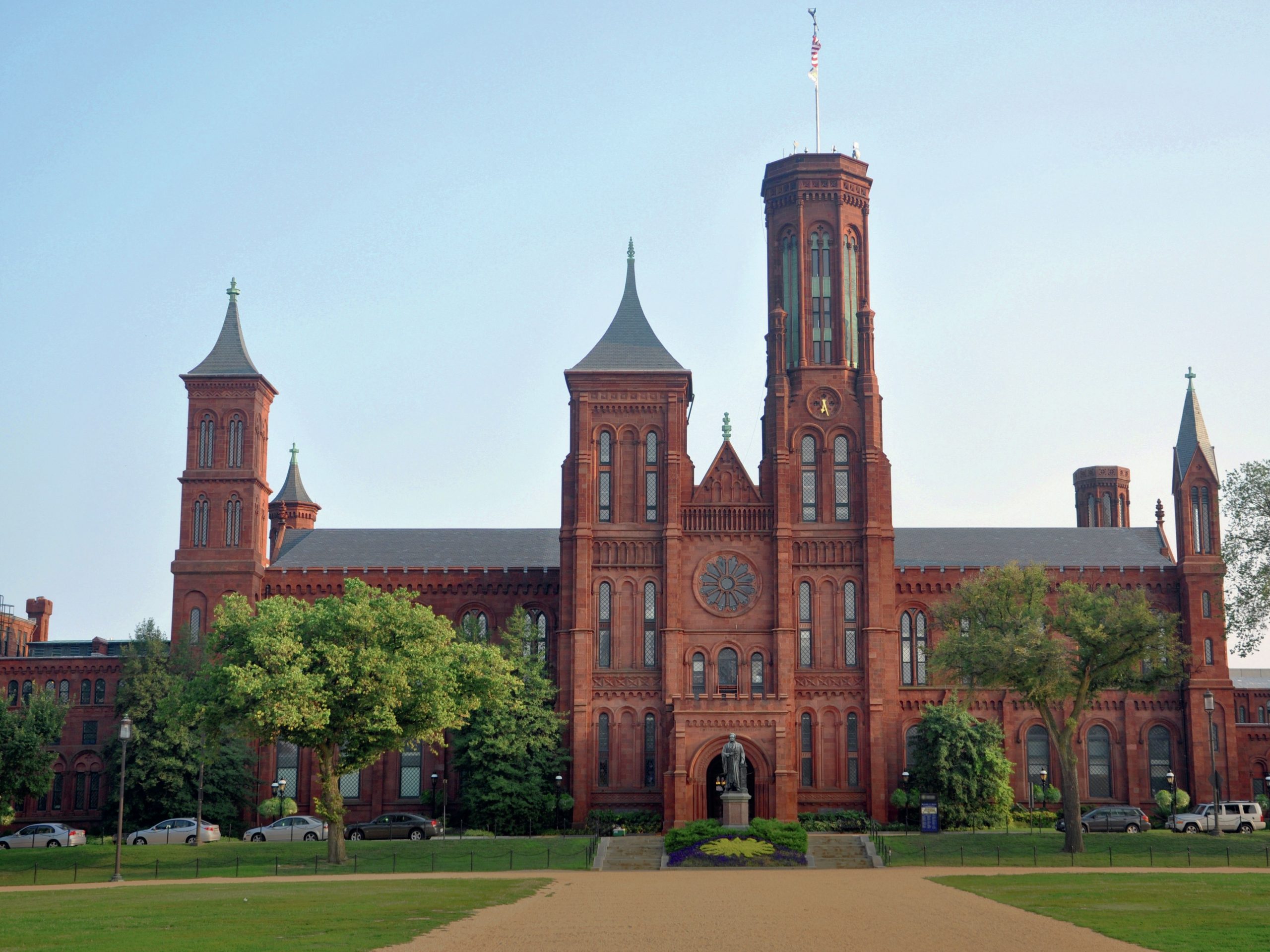A reddish-coloured building with towers, with grass and trees in front of it.