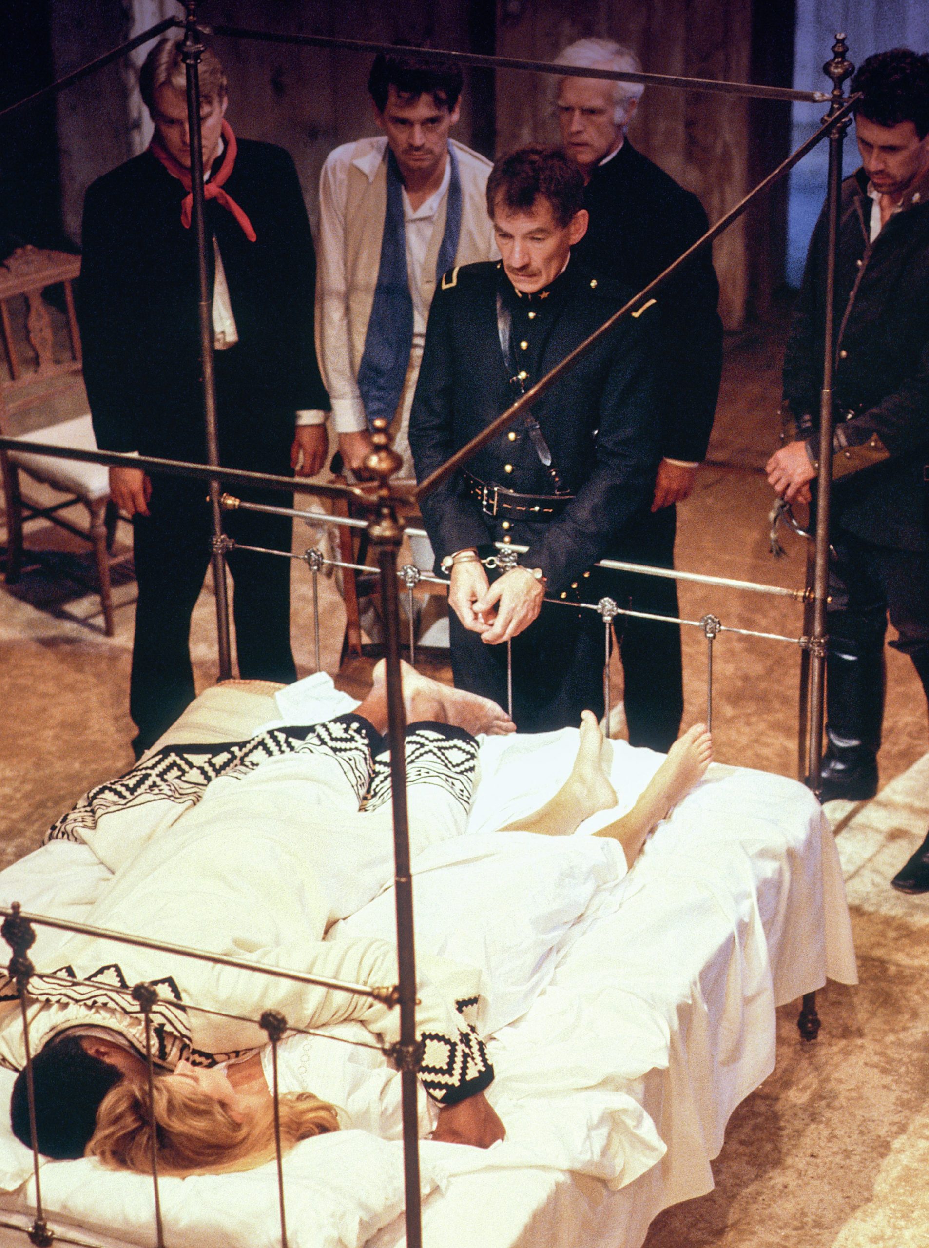 Othello and Desdemona lying on a bed on stage, watched by other characters, including a handcuffed Iago.
