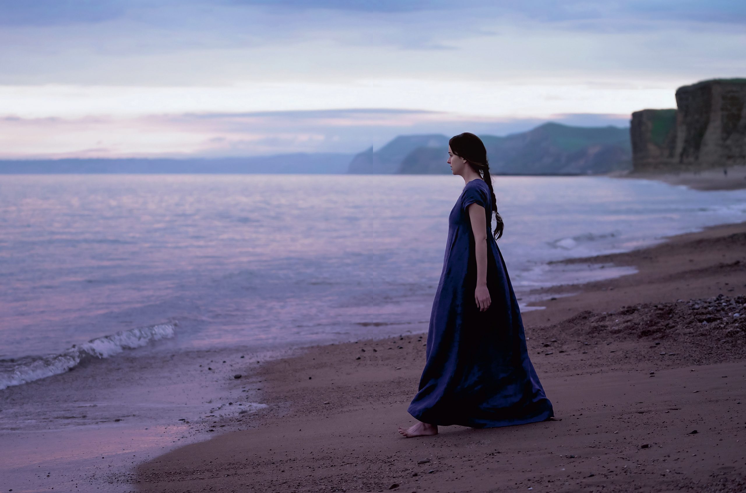 Anne Elliot walking on a beach next to the sea. She is wearing a long blue dress and is barefoot.