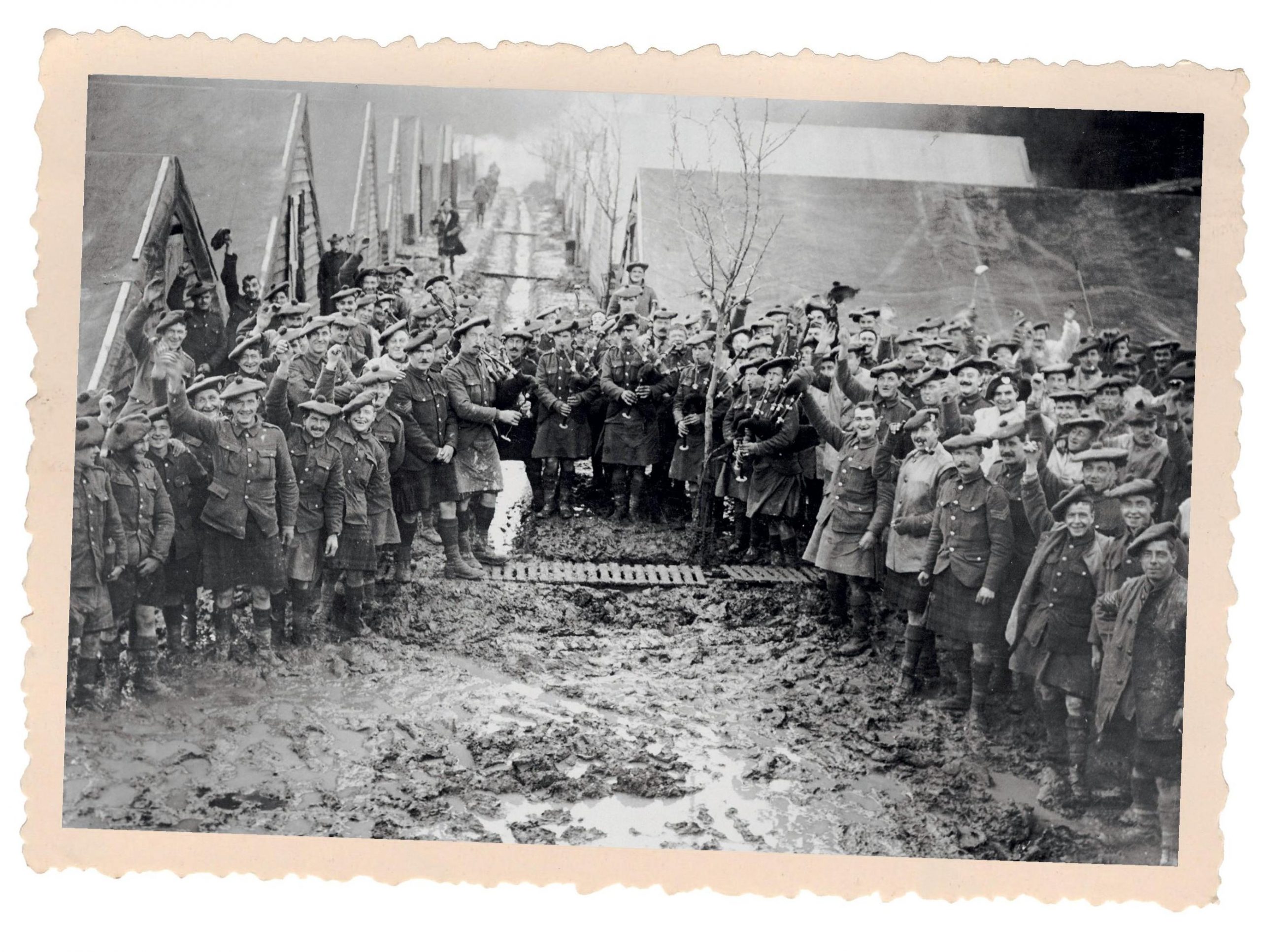 Black and white photo of around fifty soldiers, wearing tam o’ shanters. Many are smiling and waving to the camera. There are several bagpipers.