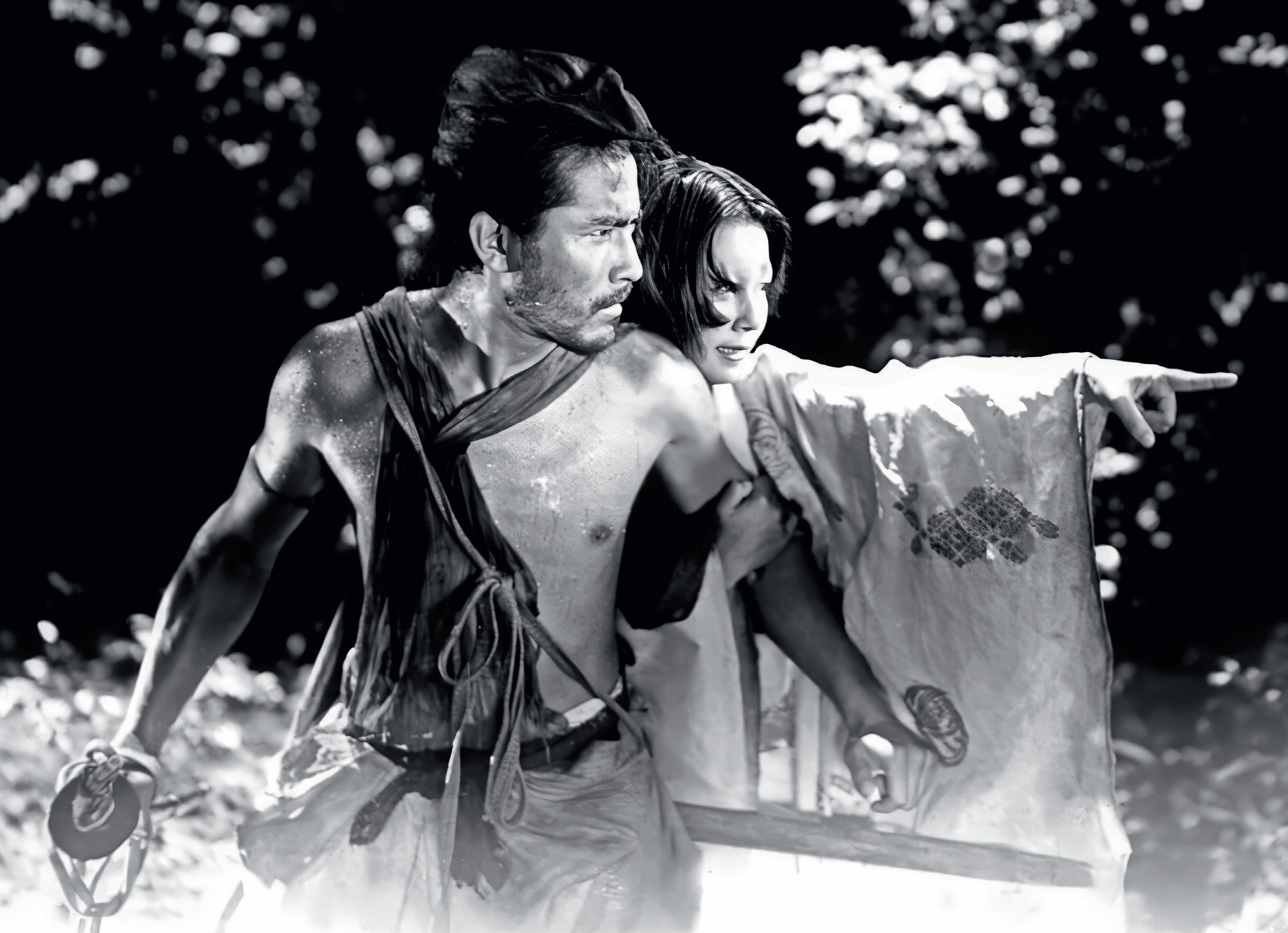Black and white photo with a Japanese man in the foreground holding a sword, and a woman behind him, pointing at something out of shot.