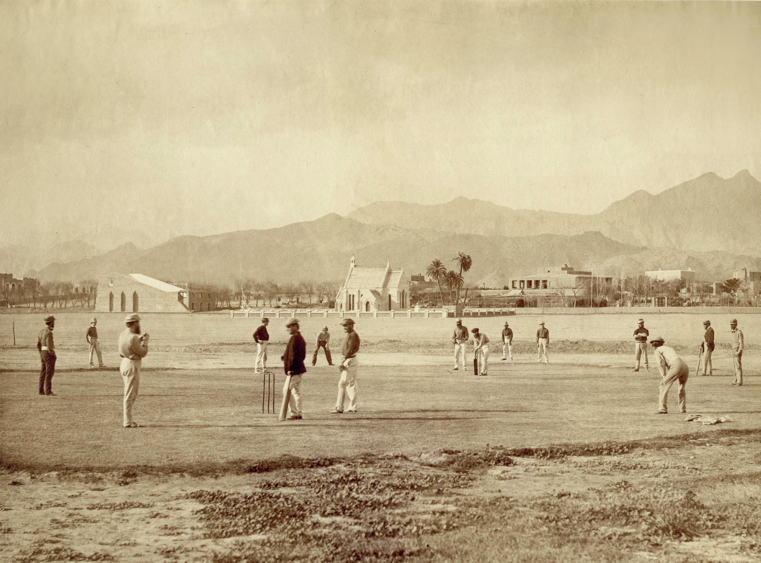 Old sepia photograph of people playing cricket in a field. There are buildings and mountains in the distance.