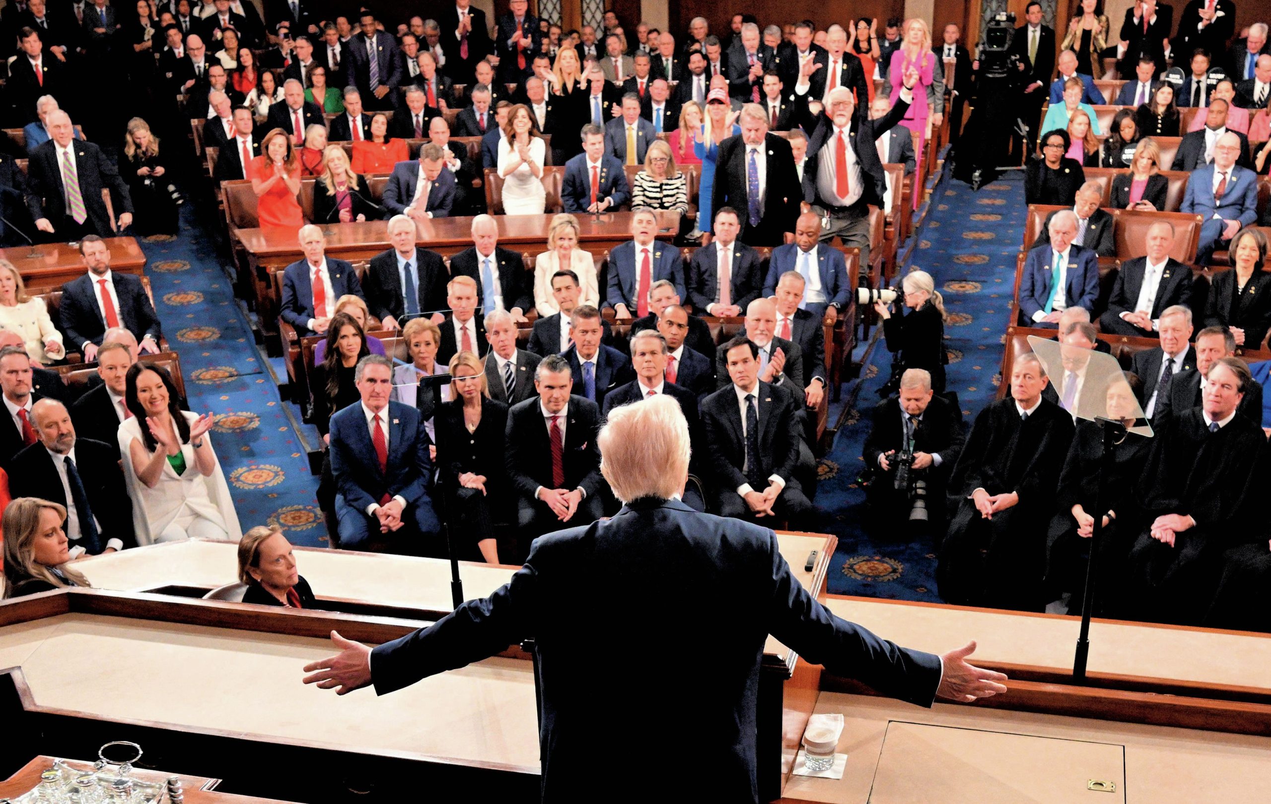 Trump shown from the back, with his arms outstretched. In front of him are about 100 members of Congress, including Supreme Court justices.