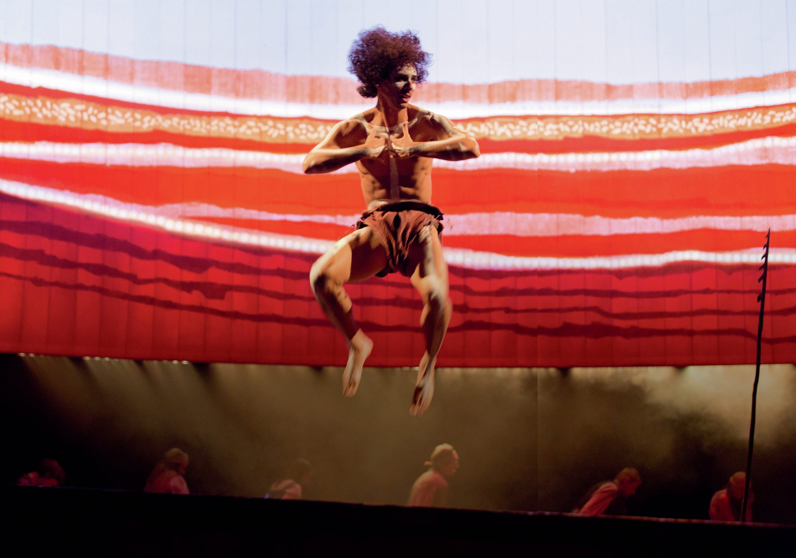 Man on stage jumping high, with other cast members behind.