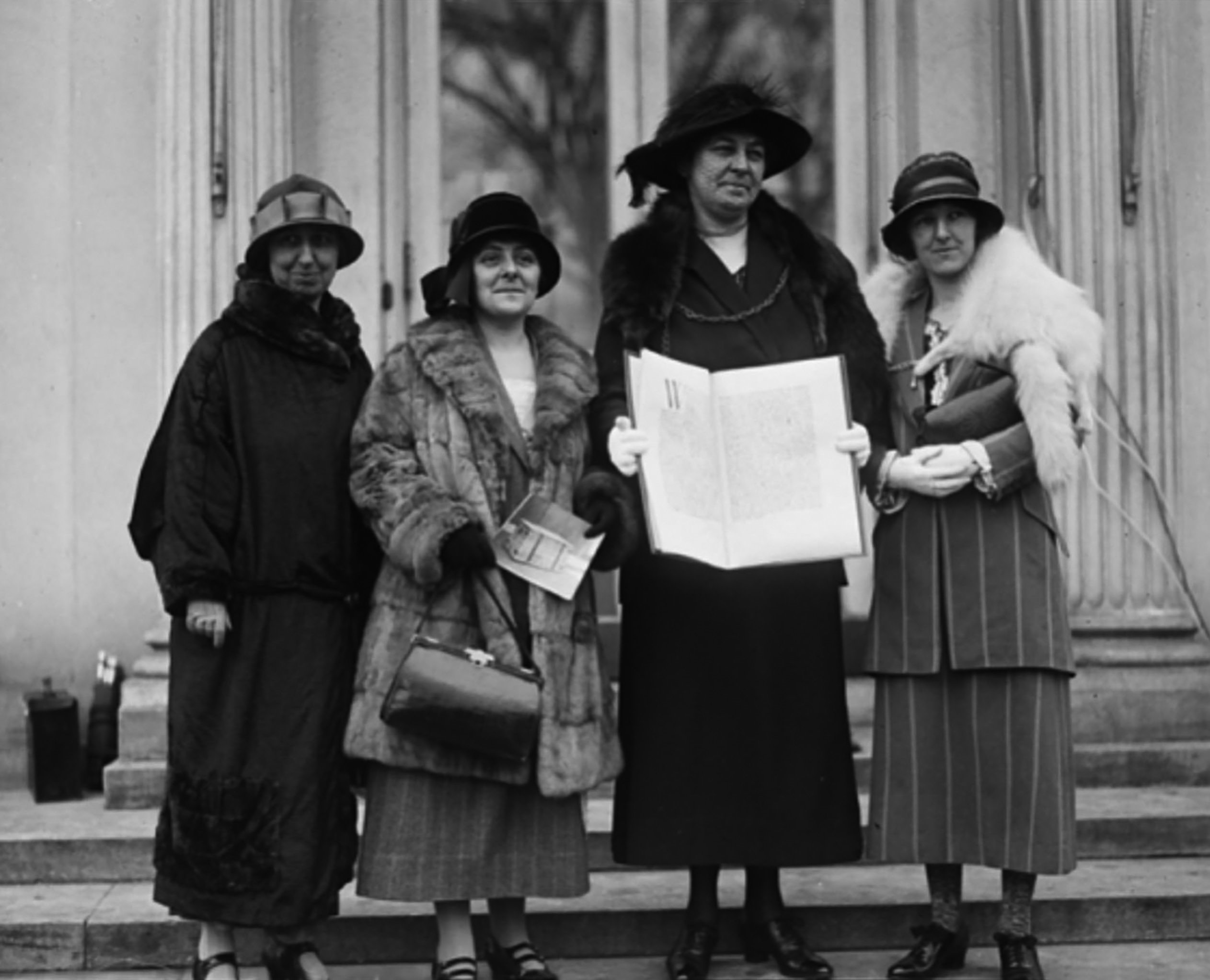 Four women stand in front of a building. One holds a petition.