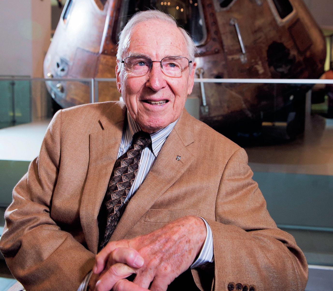 A man in a brown suit and glasses, in front of a space capsule