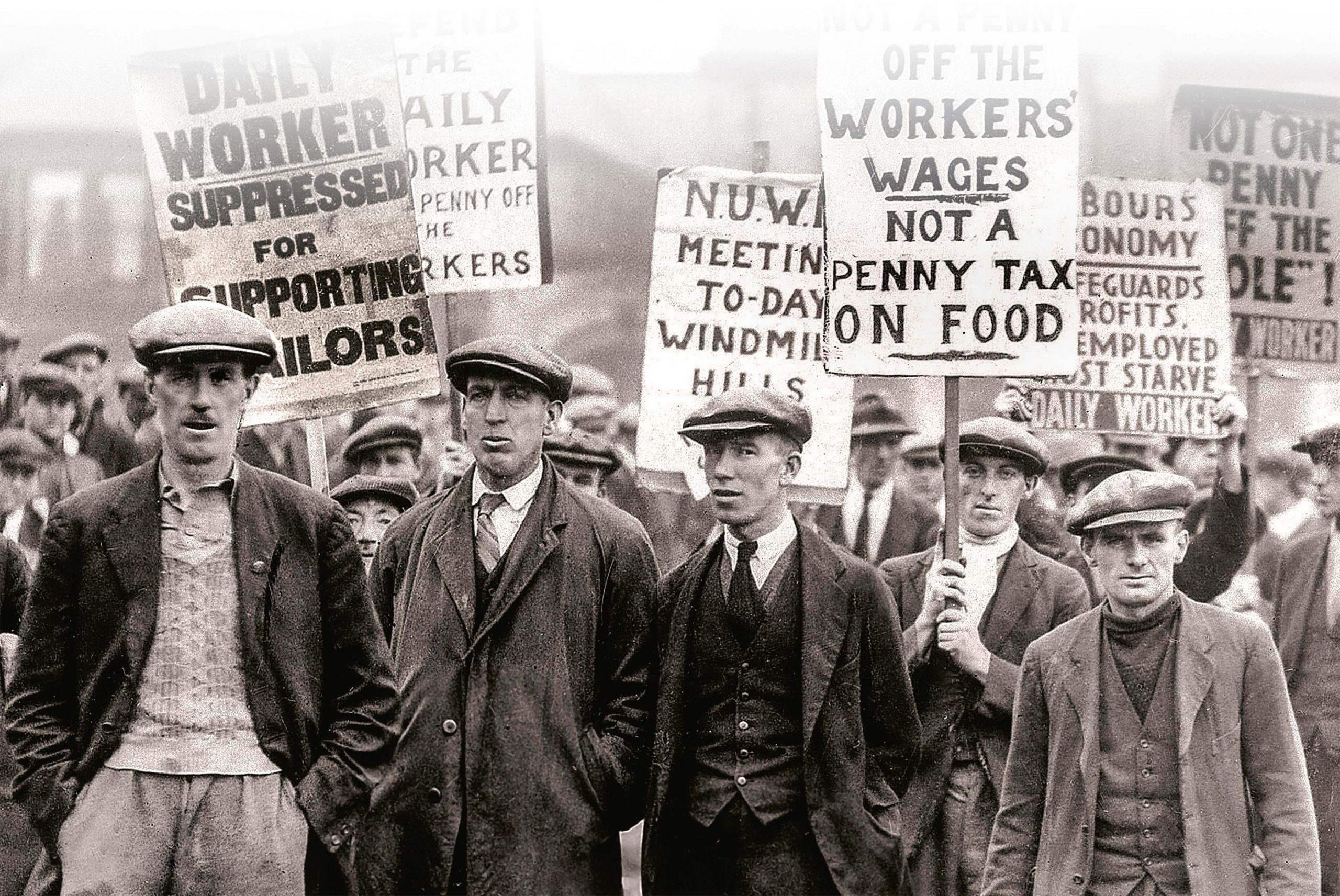 A crowd on men wearing suits and caps holding protest signs. One sign says ‘Workers’ wages. Not a penny tax on food’